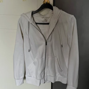Vit hoodie med dragkedja från H&M - Vit hoodie från H&M med huva, snörning och praktisk dragkedja framtill. Tröjan har två fickor och ribbade muddar vid ärmslut och nederkant. Perfekt för chill dagar och enkel att matcha med jeans eller mjukisbyxor.
