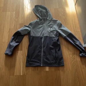 Grå vindjacka från Under Armour XS - Snygg tvåfärgad vindjacka från Under Armour i grå och mörkgrå toner. Jackan har huva, hel dragkedja framtill och lång ärm. Perfekt för dig som gillar sportig stil och vill ha något lätt och funktionellt. Logga på bröstet och praktiska fickor.