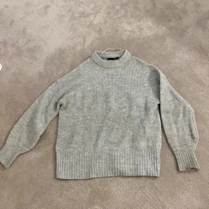 Grå stickad tröja lager 157 - Mysig grå stickad tröja från 157 med högre krage och breda ribbade muddar vid ärmslut och nederkant. Perfekt för kyliga dagar och enkel att matcha med jeans eller kjol. Tröjan har en klassisk och clean look som funkar till många stilar.