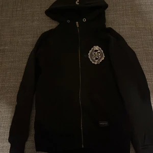 Svart hoodie från Peak Performance - Svart hoodie från Peak Performance med hel dragkedja och huva. Snyggt vitt emblem på bröstet och diskret logga på huvan. Tröjan har långa ärmar och ribbade muddar. Perfekt för chill eller träning, riktigt stilren och bekväm.