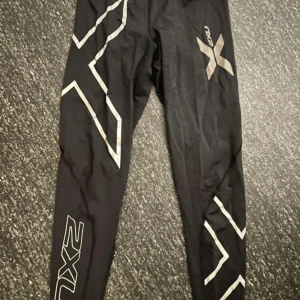 Svarta träningstights från 2XU, S - Svarta träningstights från 2XU med silvriga detaljer och logga längs benen. Tight passform och elastiskt material som passar perfekt för löpning eller gym. Snygg design med grafiska mönster och hög komfort.