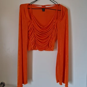 Orange croppad topp från SHEIN - Säljer en orange croppad topp från SHEIN med långa ärmar och snygg drapering framtill. Toppen har knytband i ryggen och är gjord i ett tunt, stretchigt polyestertyg. Lite genomskinlig. Katt finns i hemmet