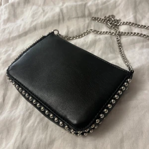  axelväska med kedja och nitar - svart axelväska i skinnimitation med silverfärgad kedjerem och coola nitar längs kanterna. 19x13x4 cm. Aldrig använd 