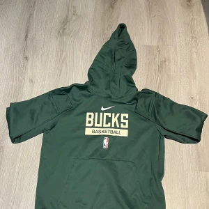 Grön Nike Bucks hoodie NBA Dri-Fit - Säljer en grön Milwaukee Bucks hoodie från Nike med Dri-Fit-material. Tröjan har huva, korta ärmar och stor Bucks-logga samt NBA-märke framtill. Perfekt för basketfans som vill ha en sportig och snygg look.
