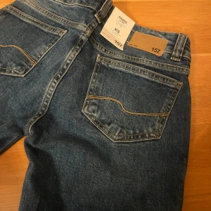 Blå bootcut jeans från 157 XS - Oanvända Snygga blå jeans från 157 i bootcut-modell med slitna detaljer på knäna. Klassisk femficksdesign, kontrastsömmar och knappar framtill. De är tyvärr för långa för mig då jag är 150 ock dessa är full length 