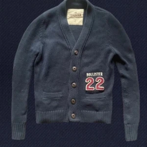 Mörkblå kofta från Hollister - Mörkblå stickad kofta från Hollister med knappar framtill och två fickor. Snygg broderad detalj med 'Hollister 22' på ena sidan. Perfekt för lager-på-lager och chill stil. Skön passform och ribbade muddar vid ärmslut och nederkant.