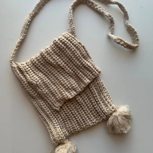 Beige stickad axelväska med tofsar - Superfin handgjord axelväska i beige, grovt stickat garn. Väskan har en lång axelrem och två stora tofsar längst ner som ger en chill och unik vibe. Perfekt för dig som gillar boho-stil och vill ha något annorlunda.