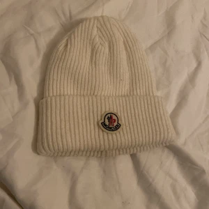Moncler mössa - Snygg vit ribbstickad mössa från Moncler med klassisk logotyp framtill. Mössan har uppvikt kant och är tillverkad i mjukt material som håller dig varm och stilren under kalla dagar. Perfekt accessoar för vinter och höst.