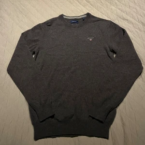 Mörkgrå stickad tröja från GANT - Snygg mörkgrå stickad tröja från GANT i superfin lammull. Rund hals med diskreta vita ränder på insidan av kragen och broderad GANT-logga på bröstet. Perfekt för dig som gillar klassisk och stilren look.