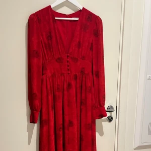 Röd långklänning från Adoore - Supersnygg röd långklänning från Adoore med blommigt mönster, v-ringning och klädda knappar framtill. Klänningen har långa ärmar med knappar vid ärmslut och markerad midja. Perfekt för dig som vill sticka ut med en elegant och trendig look.