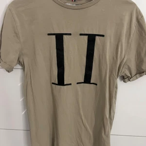 Beige t-shirt från Les Deux med tryck - Snygg beige t-shirt från Les Deux med stort svart II-tryck på bröstet. Klassisk rund hals och korta ärmar med uppvik. Mjuk bomullskvalitet som känns skön mot huden. Perfekt för dig som gillar stilrena och enkla plagg med en twist.