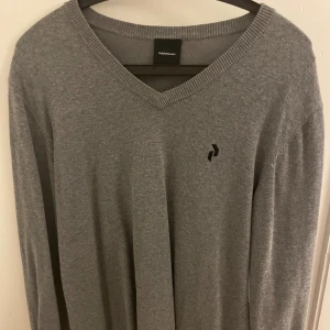 Grå v-ringad tröja Peak Performance - Snygg grå v-ringad tröja från Peak Performance med diskret svart logga på bröstet. Tröjan har ribbade muddar vid halsen och är långärmad. Perfekt för dig som gillar stilrena och klassiska plagg med en sportig touch.