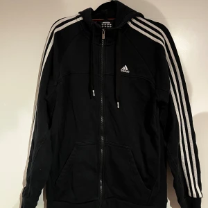 Svart Adidas hoodie med dragkedja - Svart hoodie från Adidas med klassiska vita ränder längs ärmarna och vit logga på bröstet. Tröjan har huva med snörning, dragkedja framtill och två fickor.
