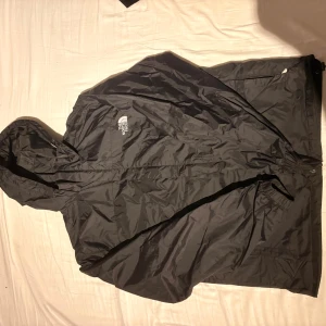 Svart vindjacka från The North Face - Svart vindjacka från The North Face med huva och dragkedja. Jackan har logga på bröstet och bak. Perfekt för blåsiga dagar och enkel att packa ner i väskan. Snygg och funktionell design med justerbar nederkant.