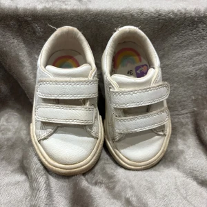 Vita sneakers från Next med kardborre - Söta vita sneakers från Next med två praktiska kardborreband och rund tå. Skorna har en platt sula och är tillverkade i syntetmaterial. Perfekta för små fötter som vill springa runt bekvämt .