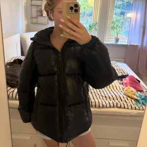 Svart pufferjacka med huva - Säljer en svart pufferjacka med huva och dragkedja framtill. Jackan är quiltad och har en lite glansig yta. Perfekt för kalla dagar och har en skön, oversized passform. Fickor på sidorna och dragsko nertill för extra komfort.