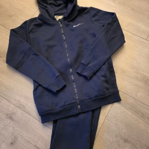 Marinblå hoodie från McKenzie XL - Mörkblå hoodie från McKenzie med dragkedja och huva. Tröjan har ribbade muddar, fickor och diskret logga på bröstet. Perfekt för chill dagar eller när du vill ha en enkel och snygg look.