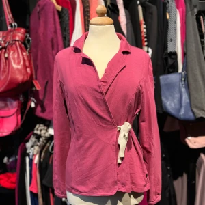 Rosa/lila omlottkofta från House of Lola - Snygg rosa/lila omlottkofta från House of Lola med vit knytning i sidan. Plagget har krage, lång ärm och är tillverkad i bomull och Elastan. Skulle säga att den är en S/M i storleken.