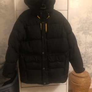 Svart pufferjacka - Säljer en svart Dunjacka med huva och snygga gula snören vid halsen. Jackan har stora fickor framtill, knappar och dragkedja. Perfekt för kalla dagar när du vill hålla dig varm och ändå se cool ut.