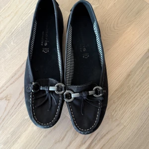 Svarta loafers i skinn med detaljer - Snygga svarta loafers från Tamaris i skinn med dekorativa metallspännen och knytdetaljer framtill. Skorna har kontrastsömmar och randigt foder på insidan för en cool touch. Perfekta för en stilren och avslappnad look.