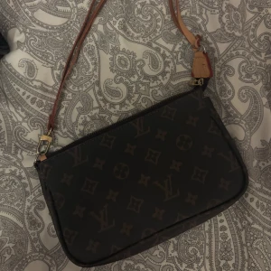 Vintage  monogram axelväska - Snygg axelväska från l v med klassiskt monogrammönster i mörkbrunt och beige. Väskan har en avlång, rektangulär form och en beige axelrem i skinn med guldfärgade metalldetaljer. Perfekt för dig som gillar lyxiga accessoarer med ikonisk design.