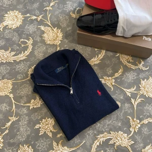 Marinblå stickad half zip från Polo Ralph Lauren - Snygg marinblå stickad tröja från Polo Ralph Lauren med half zip och röd broderad logga på bröstet. Tröjan har ribbade muddar och hög krage, perfekt för lager-på-lager. Klassisk och stilren modell som funkar året runt.