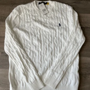 Vit kabelstickad tröja Polo Ralph Lauren - Vit kabelstickad tröja från Polo Ralph Lauren med klassisk rund halsringning och broderad marinblå logga på bröstet. Tröjan har ribbade muddar och är långärmad, perfekt för lager-på-lager under kyliga dagar.