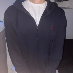 Mörkblå hoodie från Polo Sport - Svart hoodie från Polo Sport Ralph Lauren med röd logga på bröstet. Tröjan har huva med snörning, dragkedja framtill och långa ärmar. Tillverkad i mjuk bomull, perfekt för chill dagar. Klassisk och enkel design som funkar till allt.