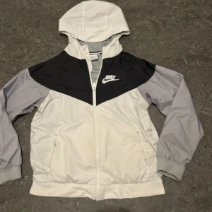 Nike vindjacka i svart, grå och vit - Nike vindjacka med huva i svart, grå och vit. Jackan har dragkedja framtill, två fickor med dragkedja och meshfoder. Klassisk Nike-logga på bröstet och sportig design. Perfekt för dig som gillar streetwear och sportig stil.