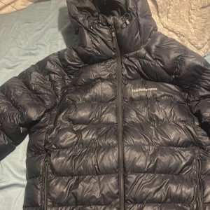 Svart Peak Performance dunjacka M - Säljer en svart dunjacka från Peak Performance i storlek M. Jackan har huva med justerbart band, två sidofickor med dragkedja och Peak Performance-logga på bröstet. Perfekt för kalla dagar med sin puffiga design och snygga look.
