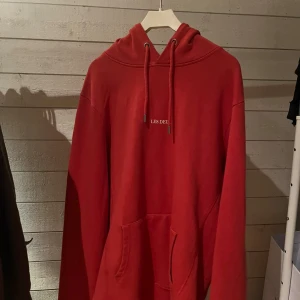Röd hoodie från Les Deux med huva - Säljer en röd hoodie från Les Deux med klassisk huva och snörning. Tröjan har en stor magficka och diskret vit logotyp på bröstet. Perfekt för dig som gillar enkel och stilren streetwear. Långärmad och i mjuk bomull för skön komfort.