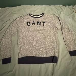 Grå sweatshirt från GANT XL - Snygg gråmelerad sweatshirt från GANT i storlek XL. Tröjan har mörkblå ribbade muddar vid hals, ärmslut och nederkant samt GANT-logga i mörkblått på bröstet. Perfekt för chill dagar och enkel att matcha med jeans eller joggers.