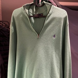 Grön half zip tröja från Polo Ralph Lauren - Snygg grön långärmad tröja från Polo Ralph Lauren med half zip och diskret lila logga broderad på bröstet. Tröjan är stickad i bomull och har en klassisk, avslappnad passform. Perfekt för dig som gillar stilrena och bekväma plagg.