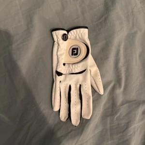 Vit golfhandske från FootJoy JL - Snygg vit golfhandske från FootJoy i storlek JL. Handsken har svarta detaljer och klassisk design med perforeringar för ventilation. Tillverkad i mjukt skinn för bra grepp och komfort. Perfekt för dig som vill ha stil och funktion på golfbanan. Vänster handske. Sitter på höger hand. För små händer eller barn.
