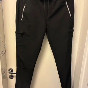 Svarta cargopants från Technicals M - Svarta cargopants från Technicals med elastisk midja och snörning. Byxorna har flera fickor med dragkedja, inklusive sidofickor och benfickor. Materialet är syntet och designen är loose fit, perfekt för en aktiv livsstil.