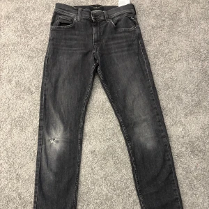 Ripped Replay jeans, slim fit passform - Snygga svarta jeans från Replay med slim fit passform. Jeansen har klassisk femficksdesign, diskret slitning på ena benet och svarta sömmar. Perfekta för dig som gillar en smal siluett och vill ha ett par stilrena jeans till vardags.