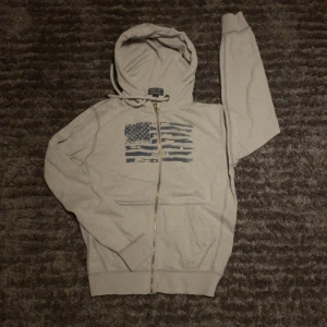 Grå hoodie Polo Jeans Company M/S - Grå hoodie från Polo Jeans Company med dragkedja och huva. Framsidan har ett tryck av en amerikansk flagga i blått och vitt. Tröjan har känguruficka och snörning i huvan. Perfekt för chill dagar och streetstyle.