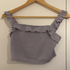 Ljuslila croppad linnetopp med volang - Superfin ljuslila croppad linnetopp med volangdetalj längs axelbanden och över bysten. Toppen har en smockad struktur och breda axelband med rynk. Perfekt för varma dagar och en söt touch till din outfit.