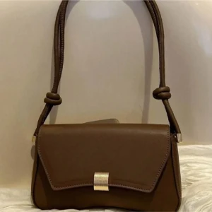 Brun axelväska med gulddetalj - Snygg brun axelväska i skinnimitation med stilren design och guldfärgat spänne framtill. Väskan har en unik form och axelremmen är dekorerad med knutar och gulddetaljer. Perfekt för dig som vill ha en trendig och praktisk väska till vardagen.