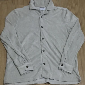 Beige frottéskjorta Studio Total M - Beige skjorta från Studio Total i mjuk frotté med klassisk krage och svarta knappar. Långärmad och loose fit, perfekt för en chill och avslappnad look. Enkel design utan mönster, passar dig som gillar stilren vibe.