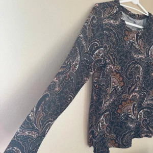 Paisleymönstrad tröja i brunt från Fransa - Skön tröja från Fransa i storlek S med ett härligt paisley-inspirerat mönster i brunt, orange, rosa, begie och svart. Tröjan har rund hals, lång ärm och mjukt material som känns skönt mot huden, stor i storleken. Det fattas en knapp på axeln, se bild 5, syns knappt men vill att ni ska veta.