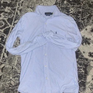 Randig skjorta Polo Ralph Lauren XS - Snygg blå- och vitrandig skjorta från Polo Ralph Lauren i klassisk oxfordstil. Skjortan har långa ärmar, knappar framtill och ikoniska broderade loggan på bröstet. Perfekt för dig som gillar stilrena och tidlösa plagg.