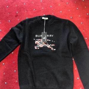 Svart stickad Burberry tröja - Svart stickad tröja från Burberry med klassiskt rutigt mönster och logga framtill. Tröjan har rund halsringning, långa ärmar och ribbade muddar. Perfekt för dig som vill ha en stilren och ikonisk look med en touch av brittisk design.