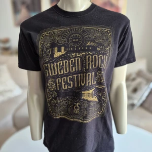 Top * Sweden Rock Festival 25 år! * Stl S unisex (dam M) - Svart T-shirt / top med tryck från Sweden Rock Festival.  * Trycket är i guld och föreställer festivalens logotyp och gitarrer.  * Baksidan har ett mindre tryck "Sweden Rock Festival" i guld. * Stl S unisex, passar dam M (Se mått) * Mått armhåla till armhåla ca 46 cm * Längd ca 64 cm * Axelsömmar ca 40 cm  * Bra skick.  * 100% bomull  Djur och rökfritt hem. Se bilder och förstora då de ingår i beskrivningen. Se gärna mina övriga annonser.