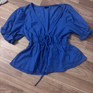 Blå omlottblus från Gina Tricot - Säljer en blå omlottblus från Gina Tricot i storlek S. Blusen har puffiga trekvartsärmar, knytband i midjan och v-ringning. Materialet är lätt och har en strukturerad känsla. Perfekt för dig som gillar en feminin och trendig look.