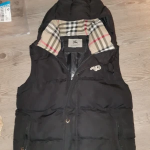 Svart dunväst från Burberry S - Svart dunväst från Burberry med klassiskt rutigt foder i beige, svart, vitt och rött. Västen har huva, dragkedja framtill och broderad Burberry-logga på bröstet. Perfekt för lager-på-lager och snygg streetstil.