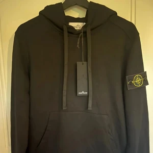 Svart hoodie från Stone Island - Svart hoodie från Stone Island med klassisk logga på ärmen. Tröjan har huva med snörning och en stor magficka framtill. Perfekt för dig som gillar streetwear och vill ha en stilren look.