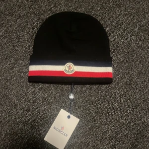 Svart Moncler mössa med ränder - Snygg svart mössa från Moncler med klassiska ränder i blått, vitt och rött vid kanten. Mössan har Moncler-logga framtill och är stickad i mjukt material som håller dig varm. Perfekt för dig som vill ha en stilren accessoar med lyxig känsla.