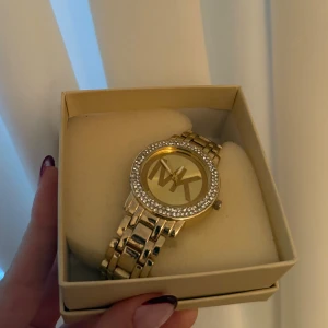 Guldig Michael Kors klocka med stenar - Säljer en snygg guldig klocka från Michael Kors med MK-logga på urtavlan. Klockan har ett armband i metall och en rund boett med glittrande stenar runt kanten. Perfekt accessoar för att lyfta din stil.