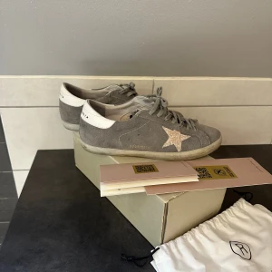 Golden Goose - Snygga sneakers från Golden Goose i grå mocka med vit häl och en råda stjärna på sidan. Skorna har snörning med tryckt logga på skosnörena och platt sula.